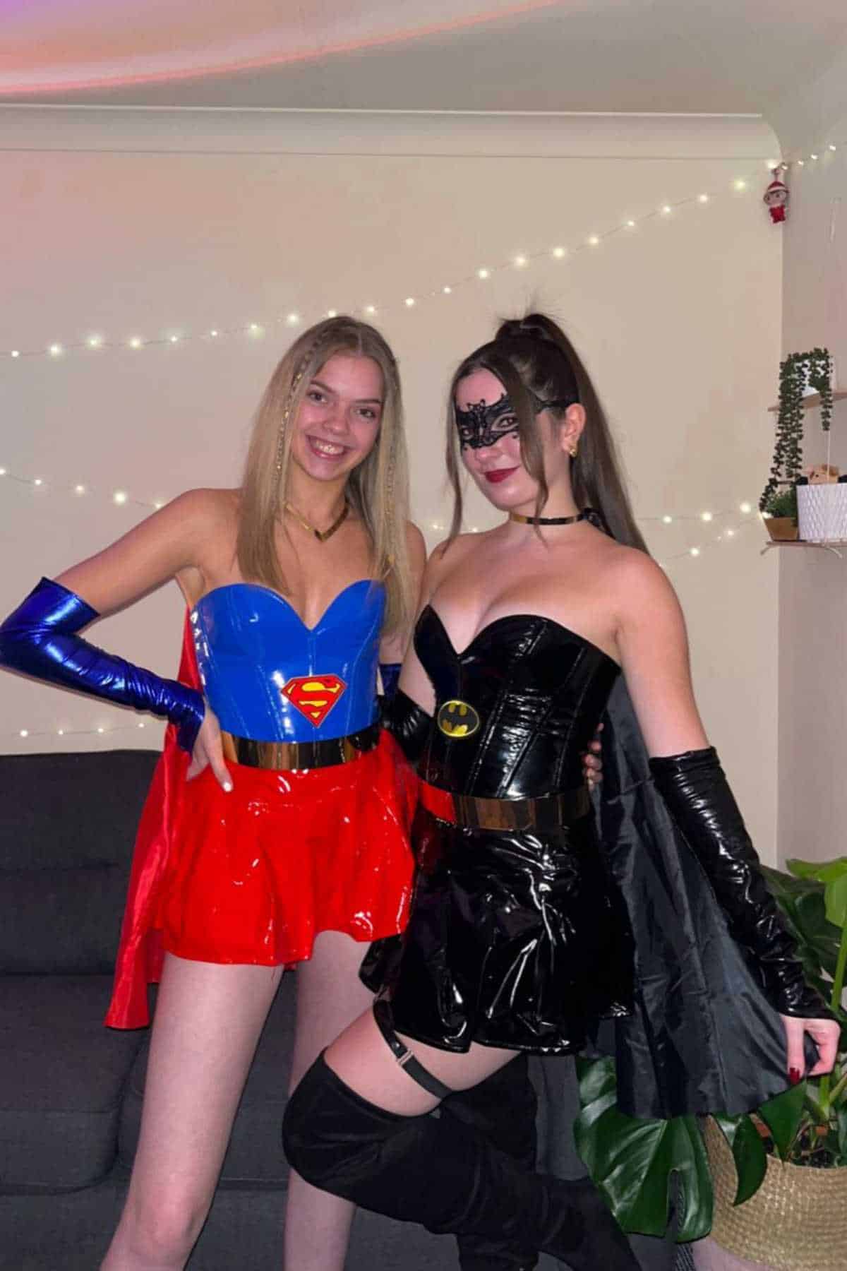 Two people are dressed in Supergirl and Batgirl costumes, standing side by side in a living room with string lights and plants in the background.
