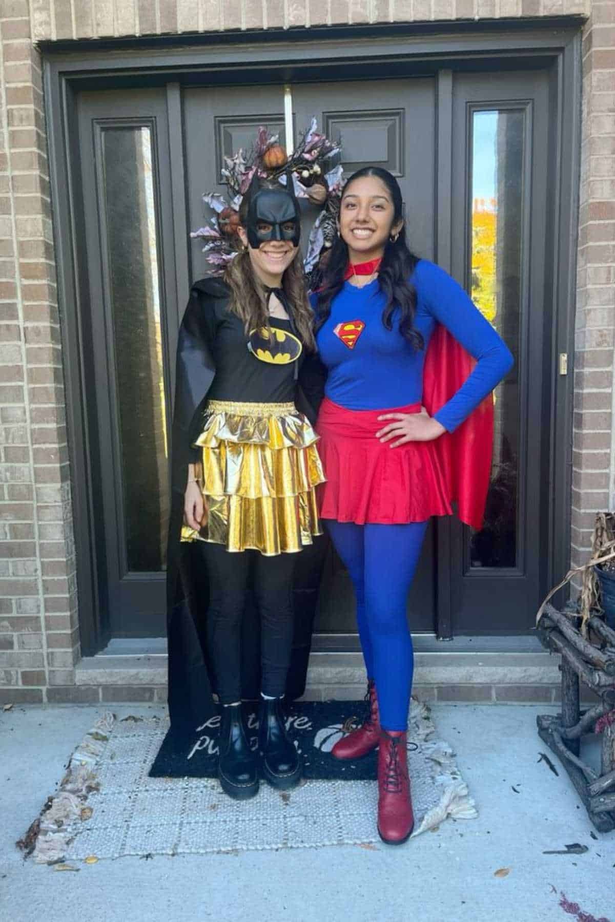 Two women stand in front of a door; one is dressed as Batgirl in black and gold, the other as Supergirl in blue and red.