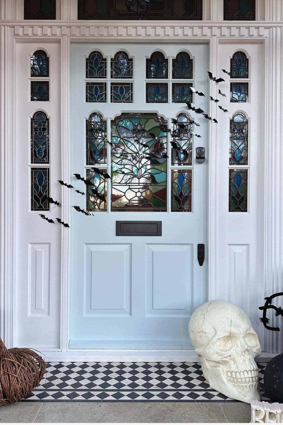 White front door with stained glass panels, black bat decorations, large skull prop, round twig decoration, and geometric black-and-white tile floor.