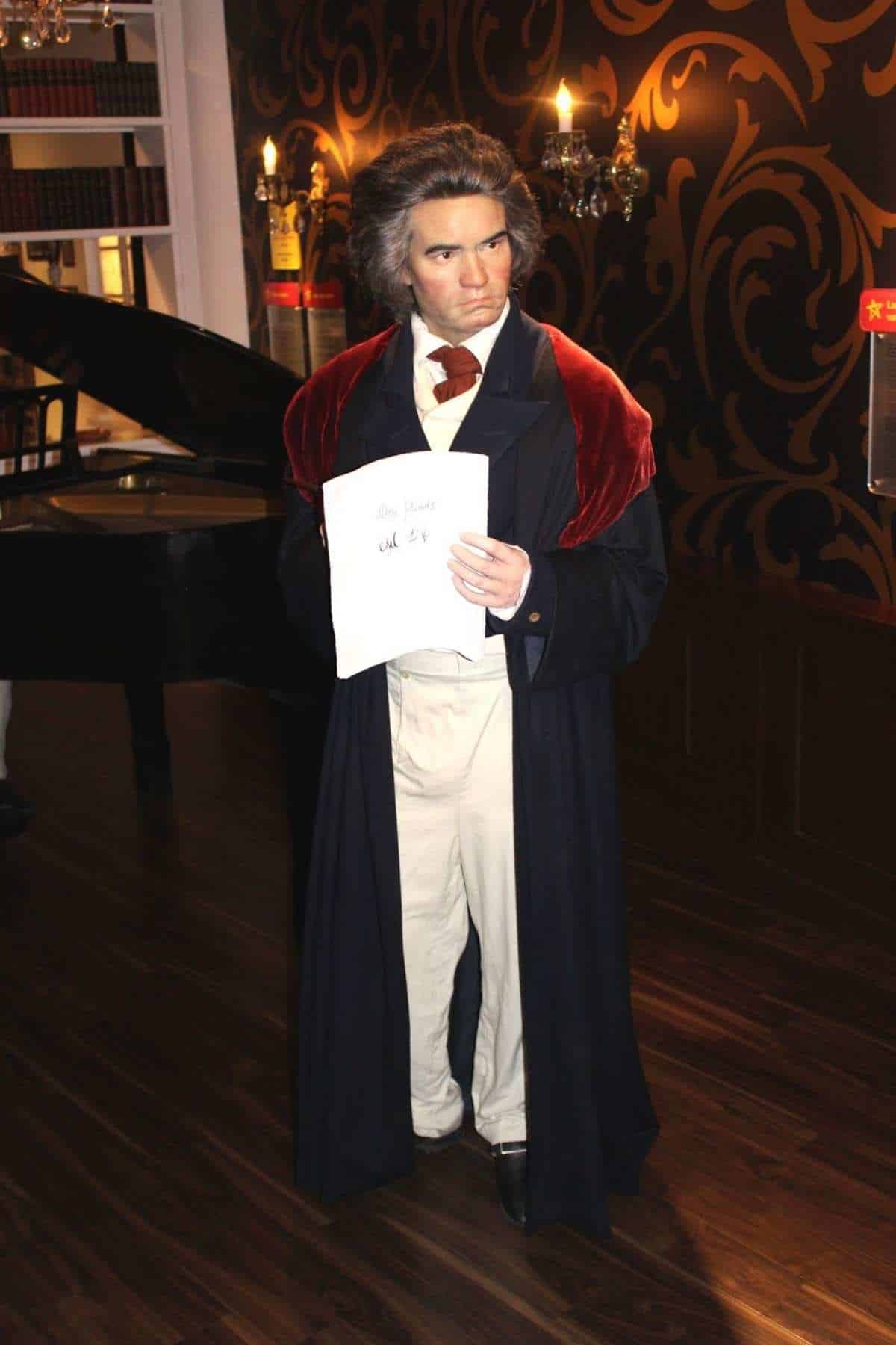 A wax figure of a man in historical attire holds a sheet of music while standing indoors next to a grand piano.