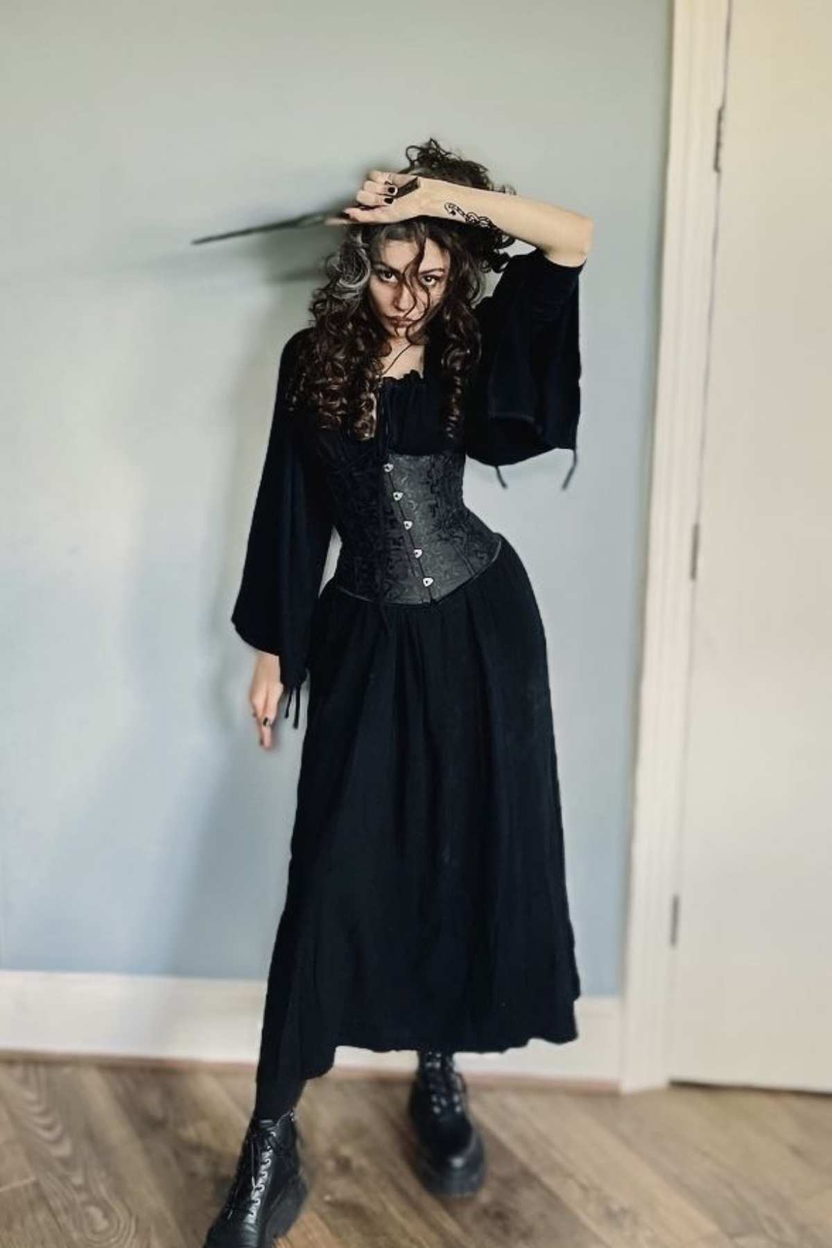 Person in a long black dress, black boots, and black corset stands indoors holding a wand, posing with one arm raised against a plain wall and wooden floor.