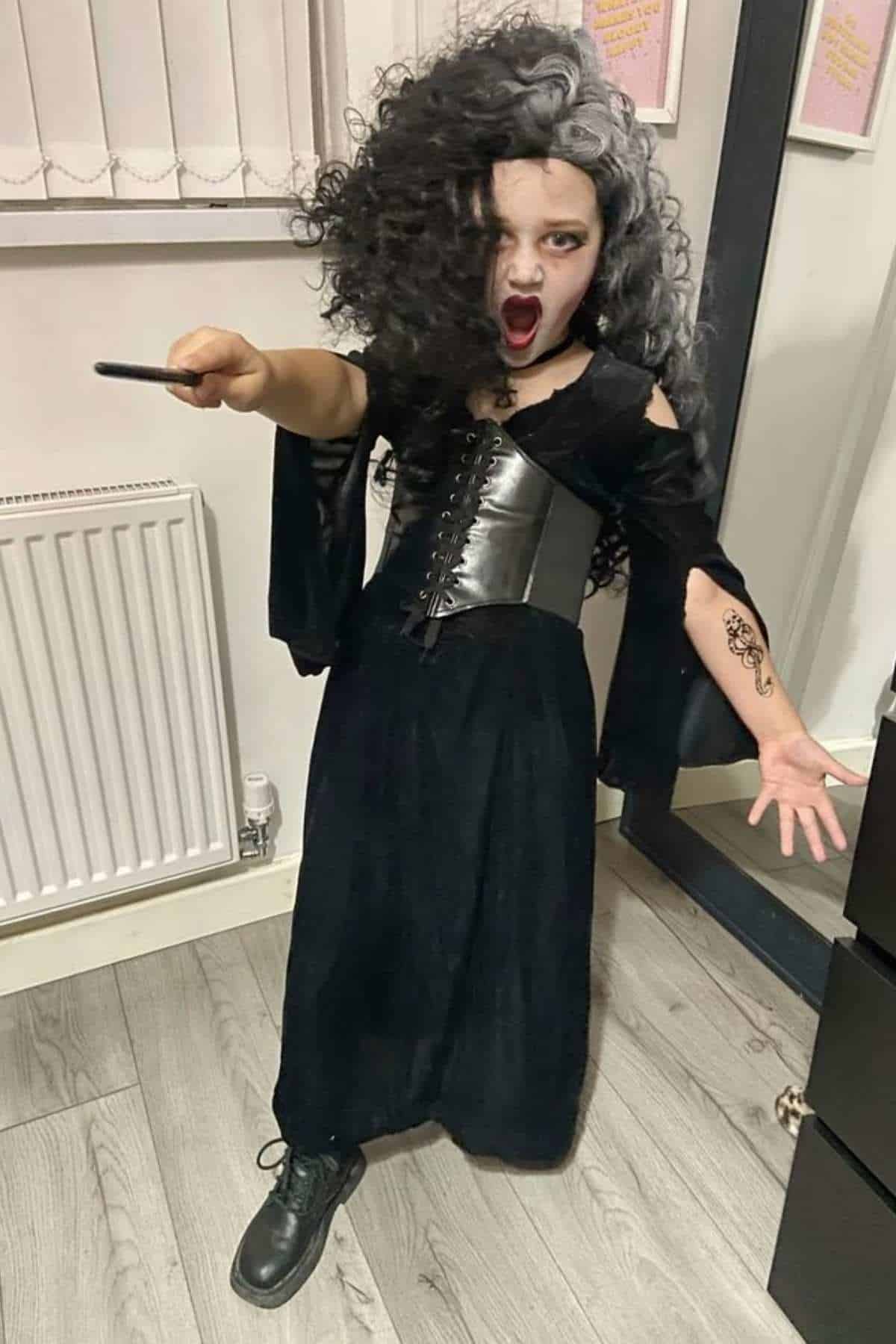 Child in black dress with curly black and gray hair, holding a wand, stands indoors with mouth open and arm extended, as if casting a spell.