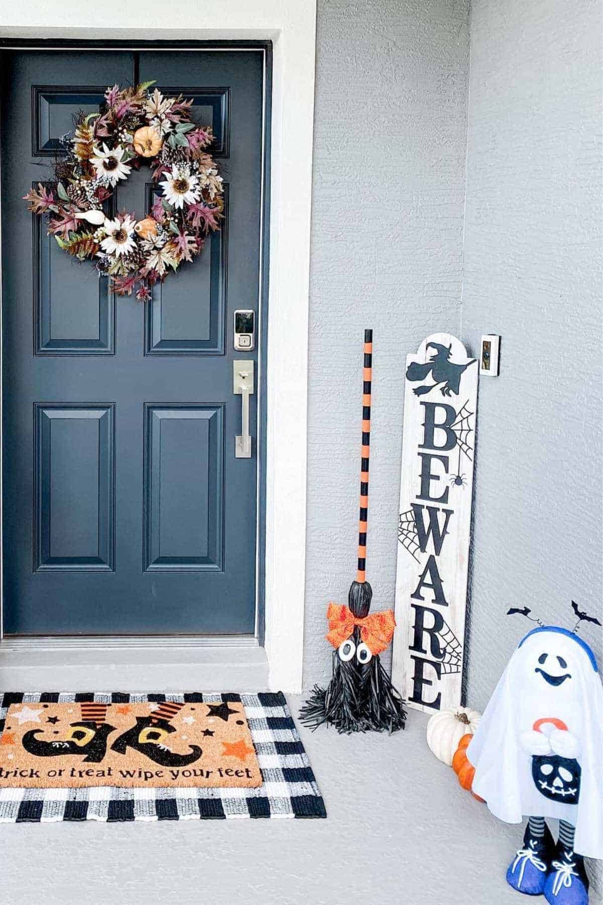A dark blue front door with a fall wreath, Halloween-themed doormat, a "BEWARE" sign, a decorative witch broom, and ghost and pumpkin decorations on a gray porch.