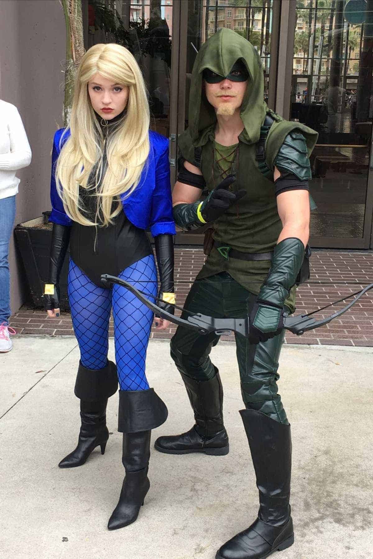 Two people in superhero costumes, one dressed as Black Canary in blue and black, the other as Green Arrow in green with a bow, pose outdoors near a building entrance.