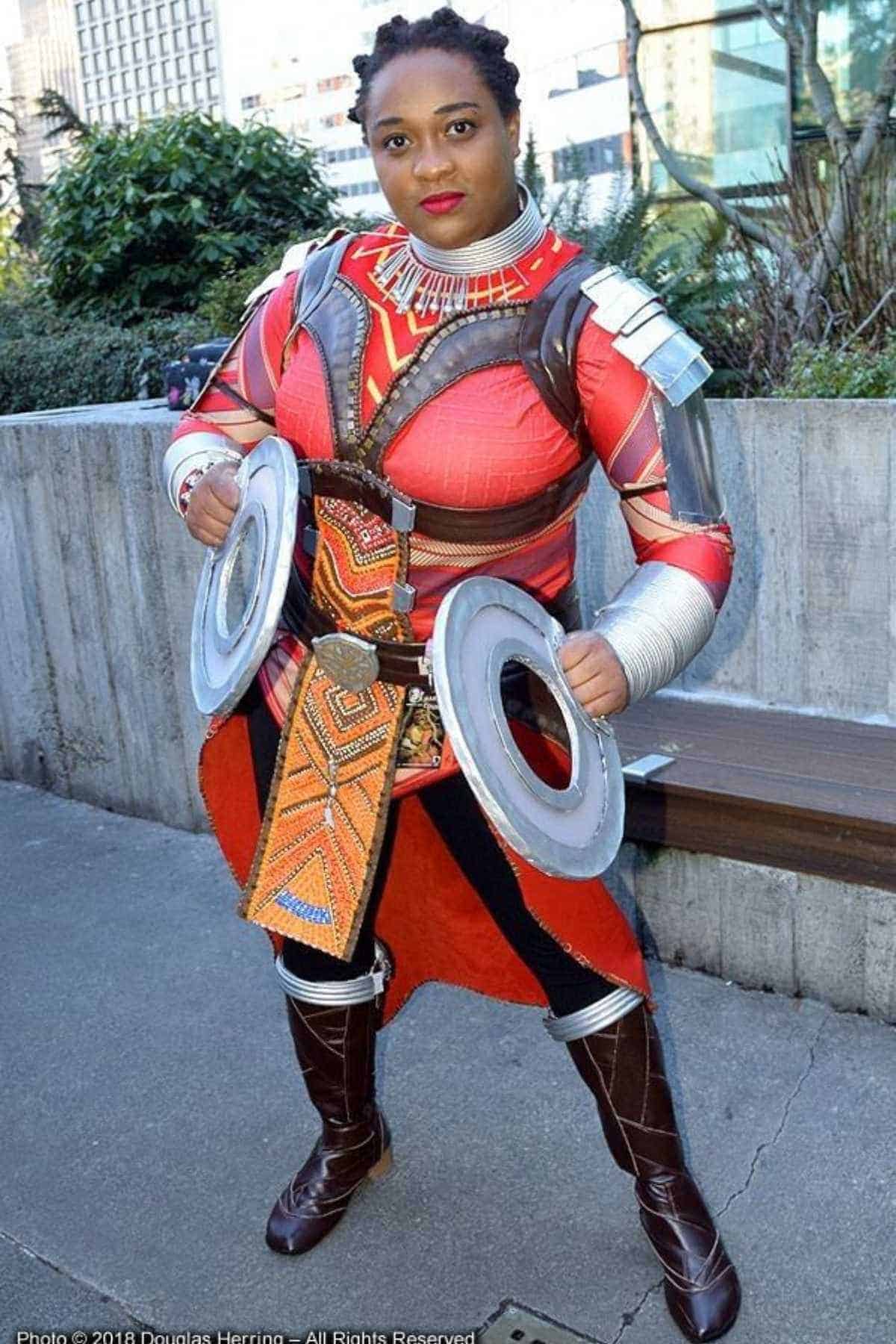 Person in detailed red and brown armor cosplay stands outdoors holding two round weapons, with buildings and greenery in the background.