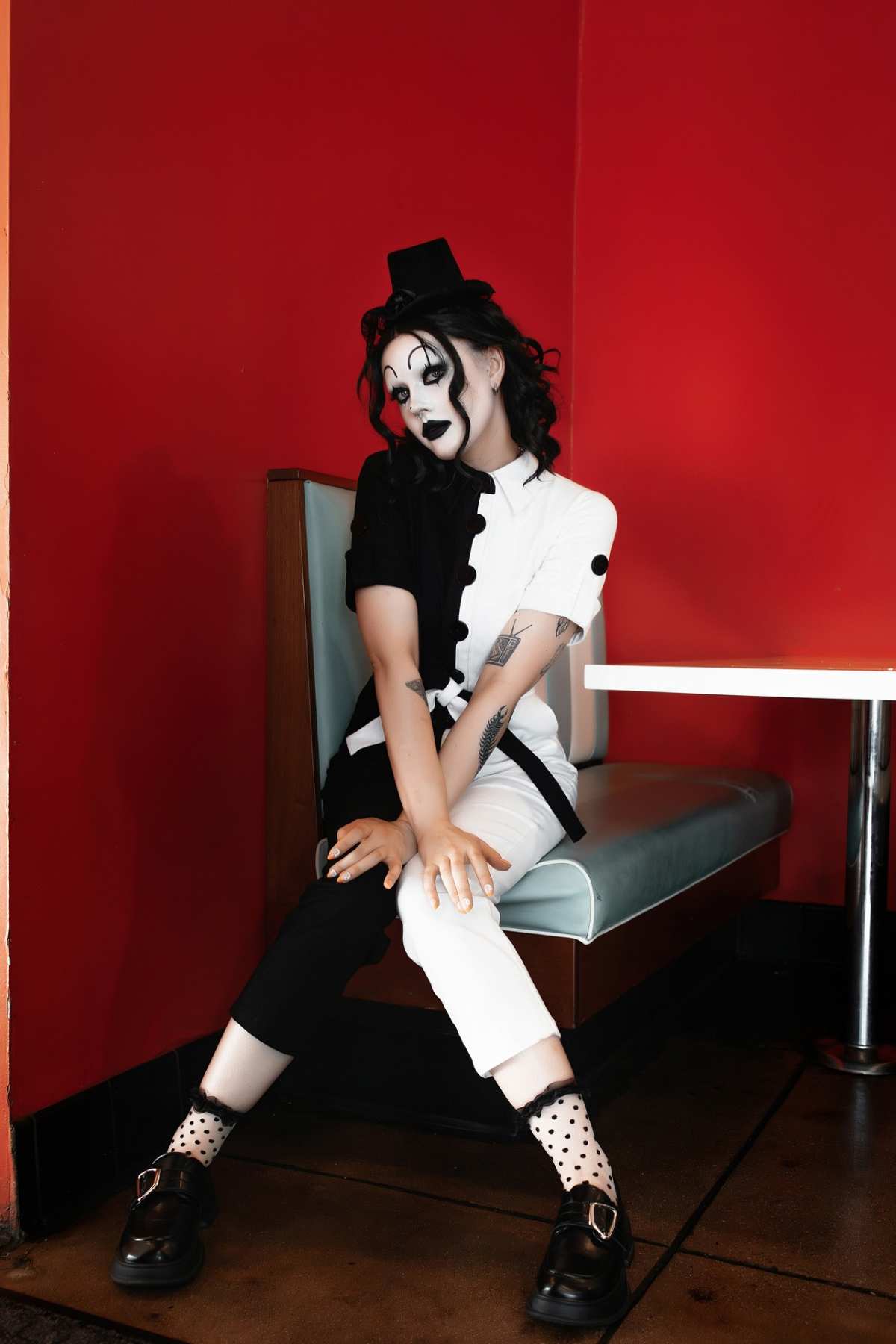 Person in a half-black, half-white mime costume with face paint sits on a booth against a bright red wall, hands resting on knees, legs apart, wearing polka-dot socks and black shoes.