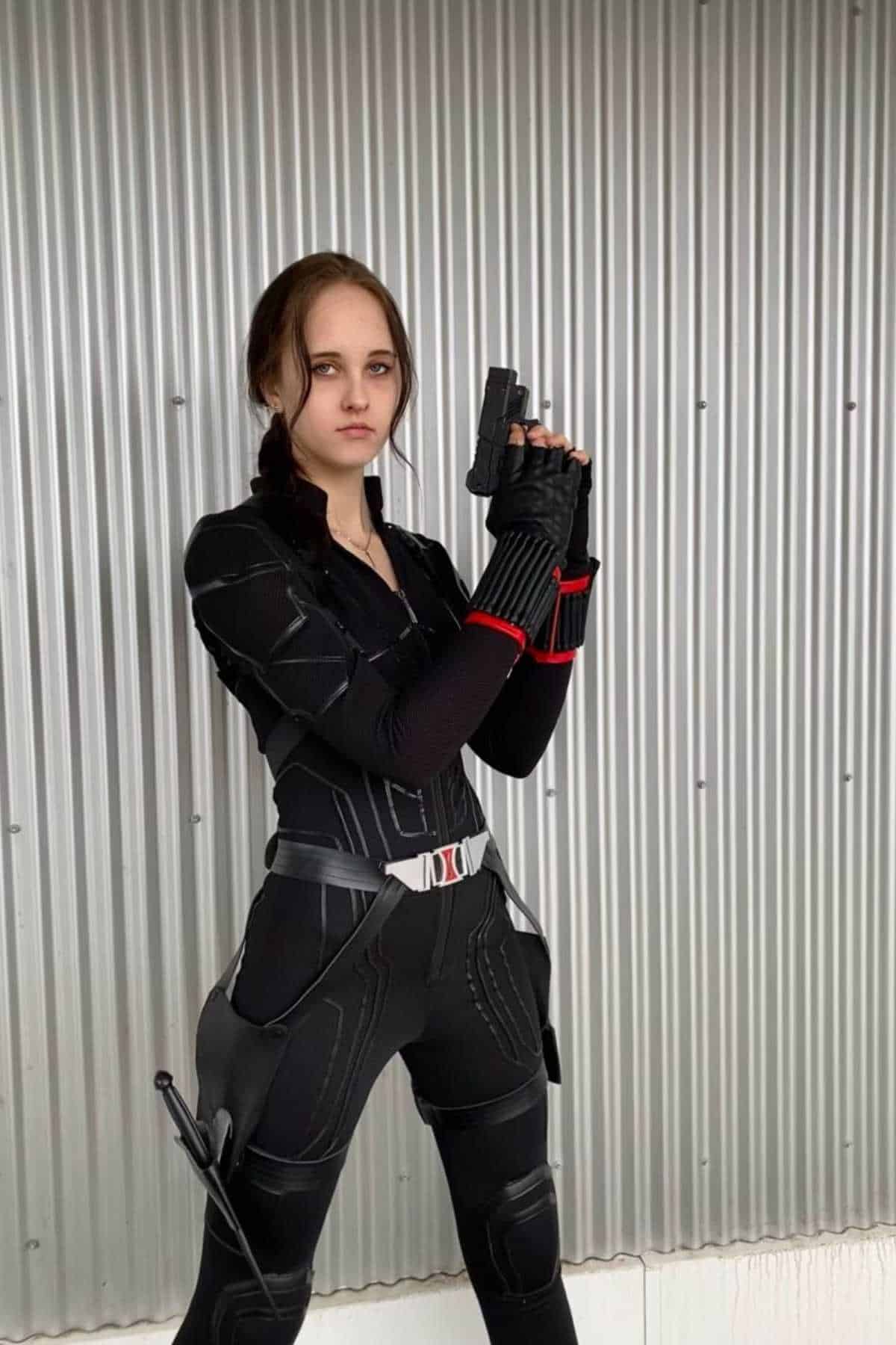 Person in a black tactical suit stands in front of a corrugated metal wall, holding a prop gun with both hands and looking directly at the camera.