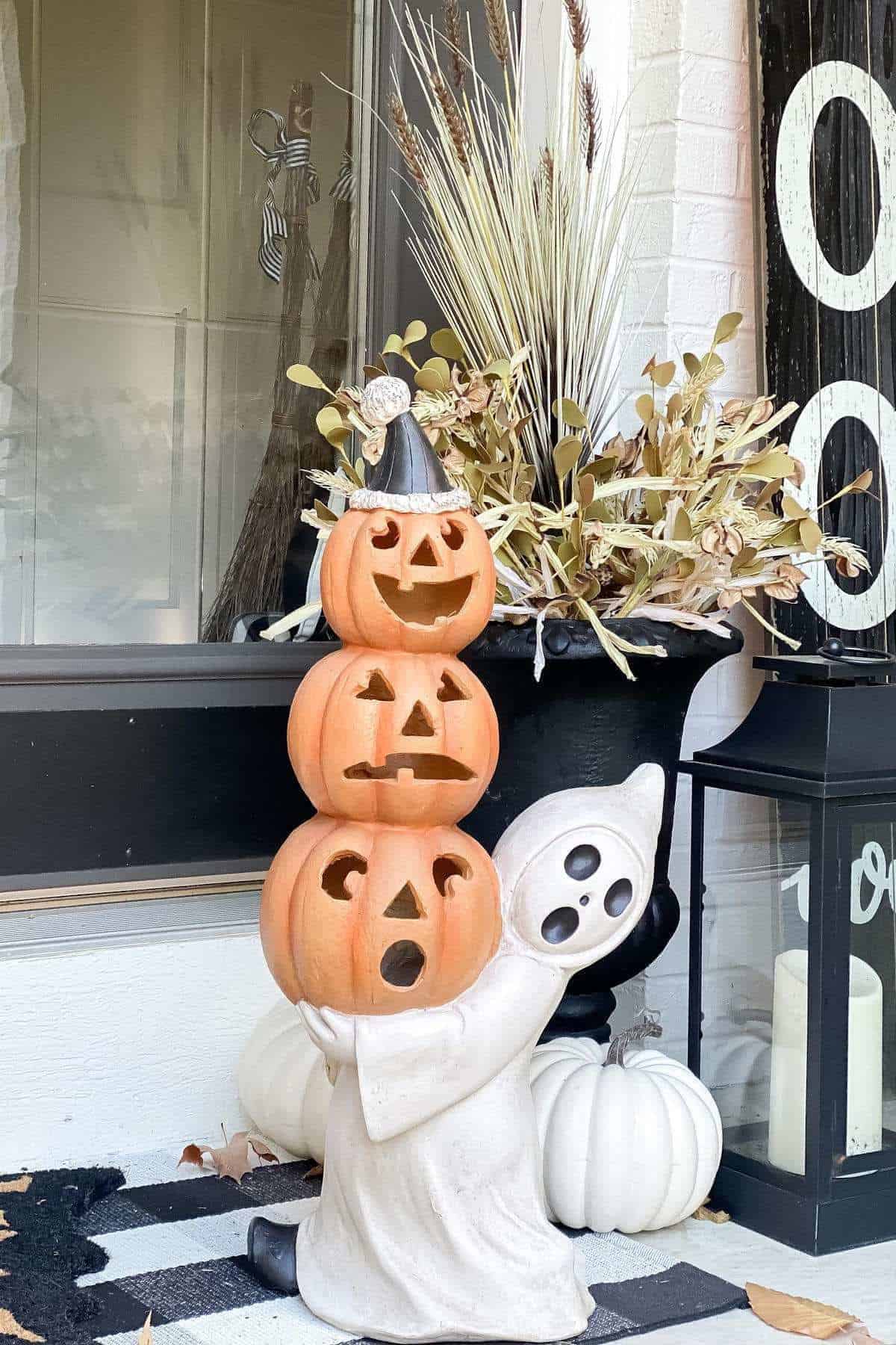 A ghost statue holds three stacked jack-o'-lanterns in front of a planter with dried grass and leaves, next to a black lantern with a white candle and a striped doormat.