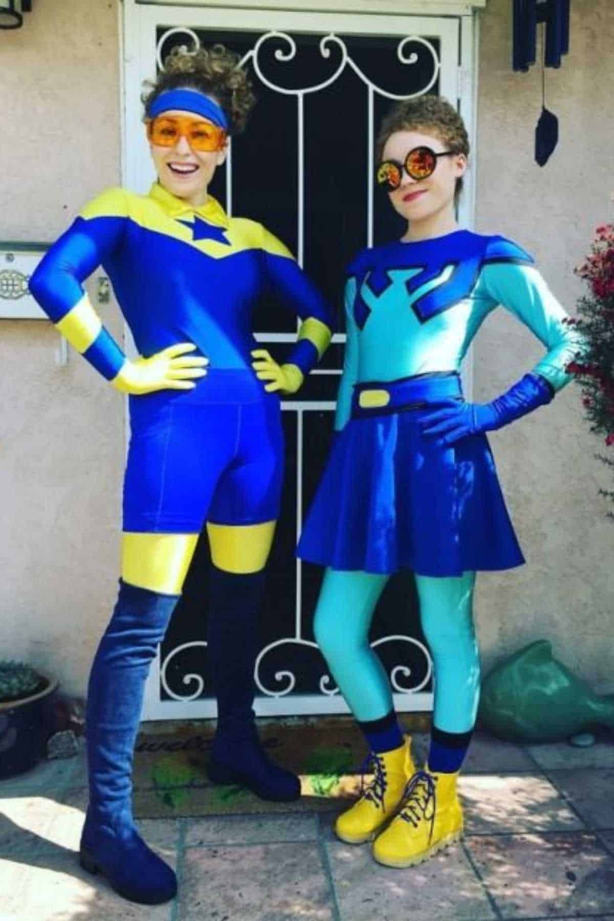 Two people wearing colorful superhero costumes pose in front of a white metal door. One wears yellow and blue, the other wears turquoise and blue with a skirt and yellow boots.