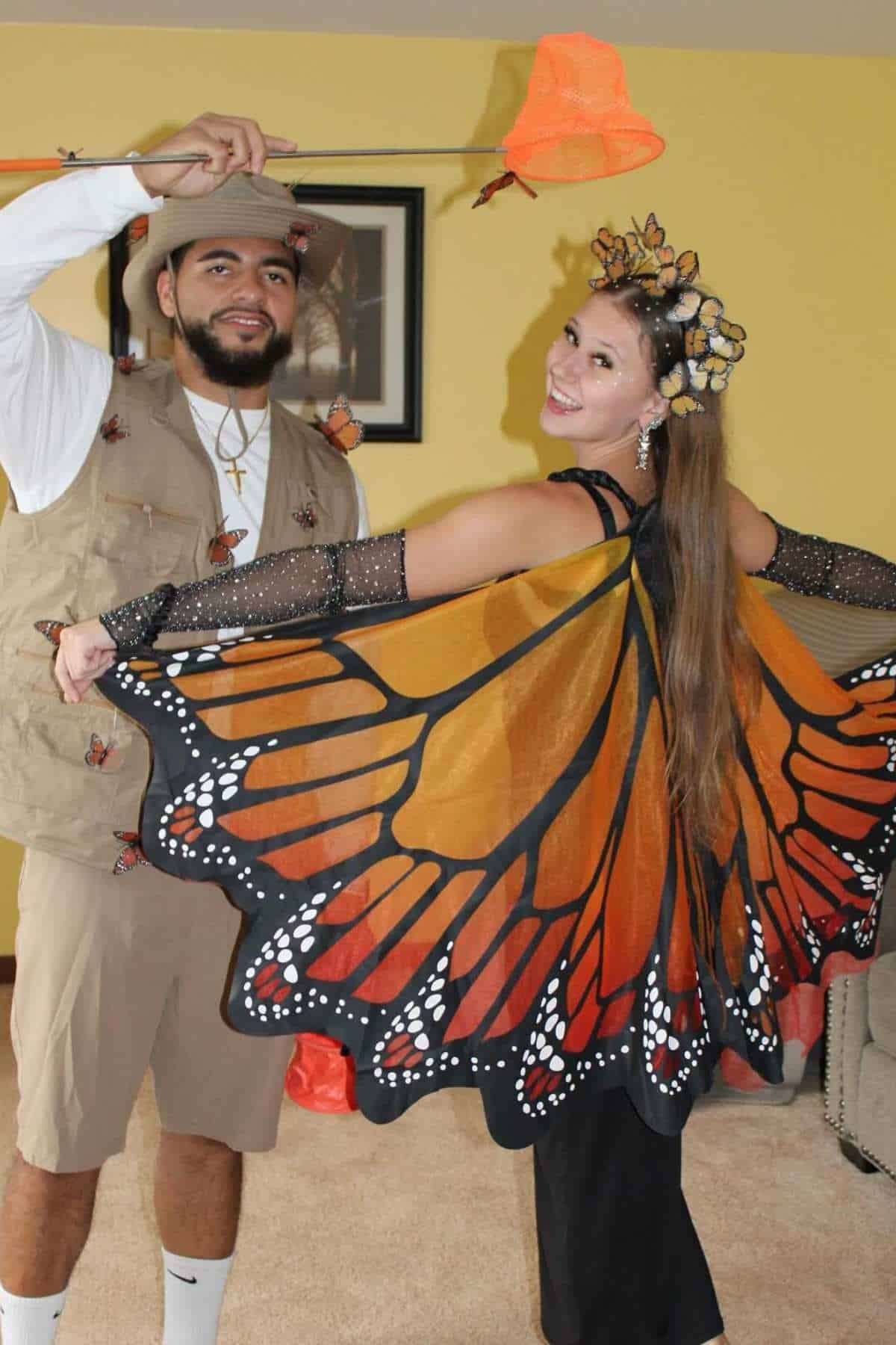 A man dressed as a butterfly catcher stands next to a woman wearing butterfly wings and a butterfly headpiece, posing indoors.