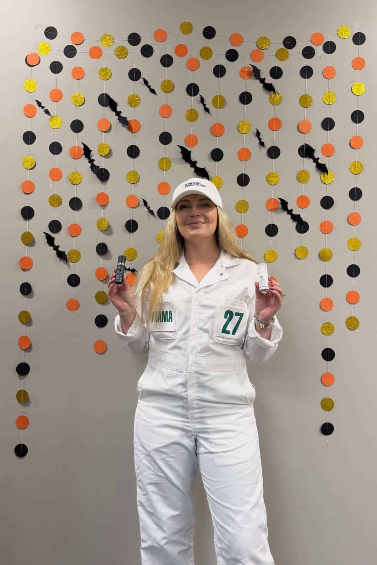 Person in a white jumpsuit and cap stands in front of a wall with orange, black, and gold dot garlands, holding objects in both hands.