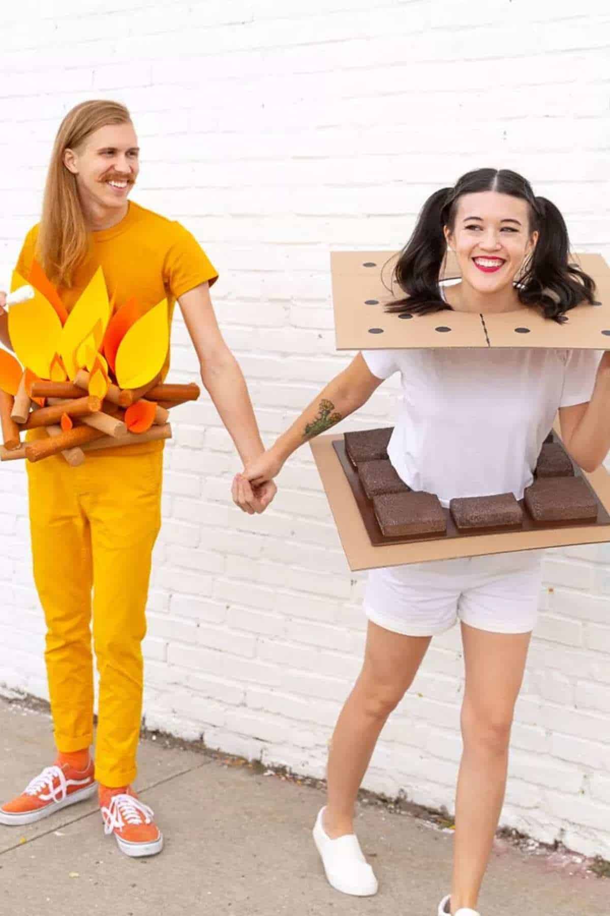 Two people holding hands, one dressed as a campfire with orange clothes and paper flames, the other dressed as a s'more with cardboard and foam props, walking by a white brick wall.