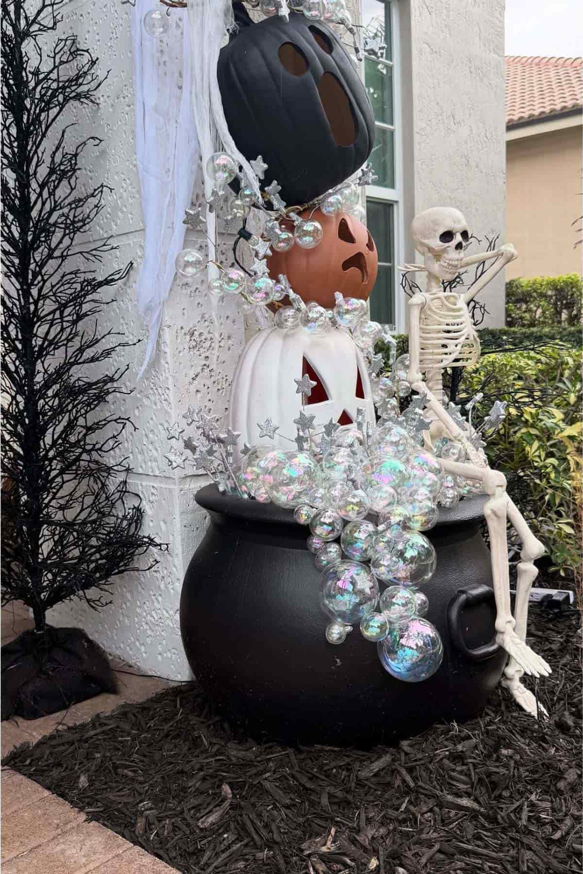 A skeleton sits on a large black cauldron with stacked jack-o'-lanterns and iridescent bubbles spilling out, set against a white wall and black tree.