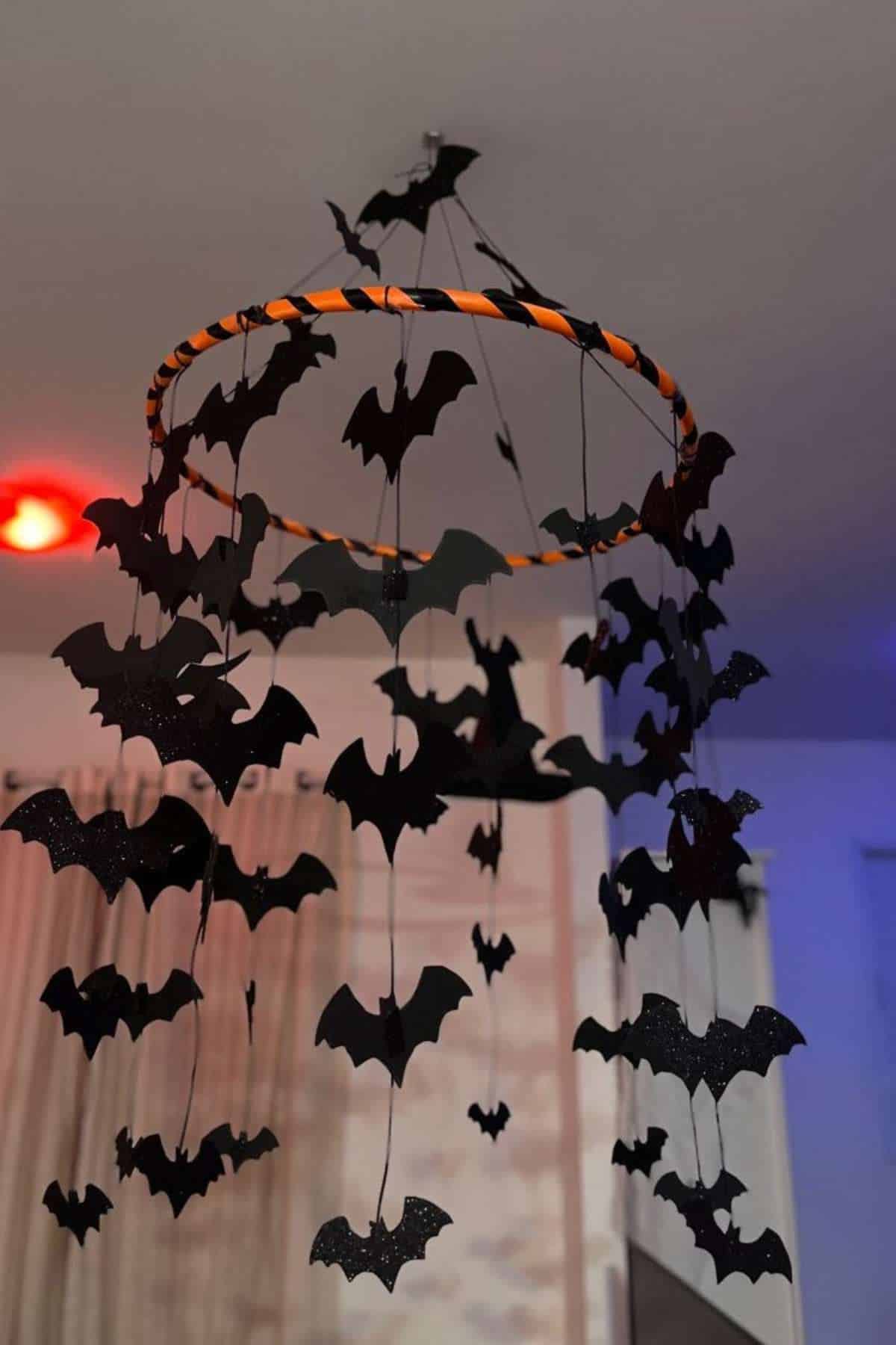 A hanging mobile made of black paper bats attached to strings and a circular frame, displayed indoors with curtains and colored lights in the background.
