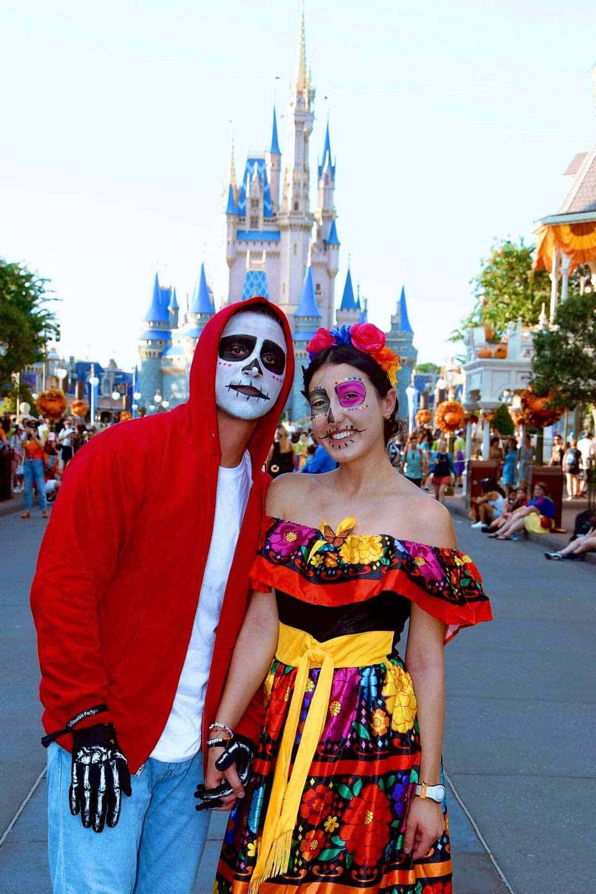 Two people in Day of the Dead-inspired costumes pose in front of a castle at a theme park, with colorful decorations and other visitors in the background.