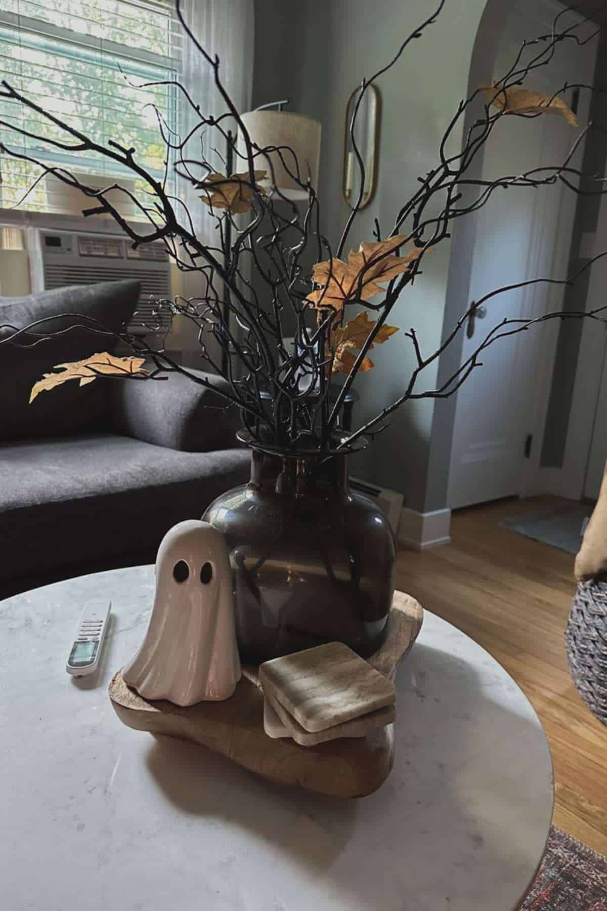 A ceramic ghost, stacked books, and a dark vase with branches and yellow leaves are arranged on a wooden tray atop a round marble table in a living room.
