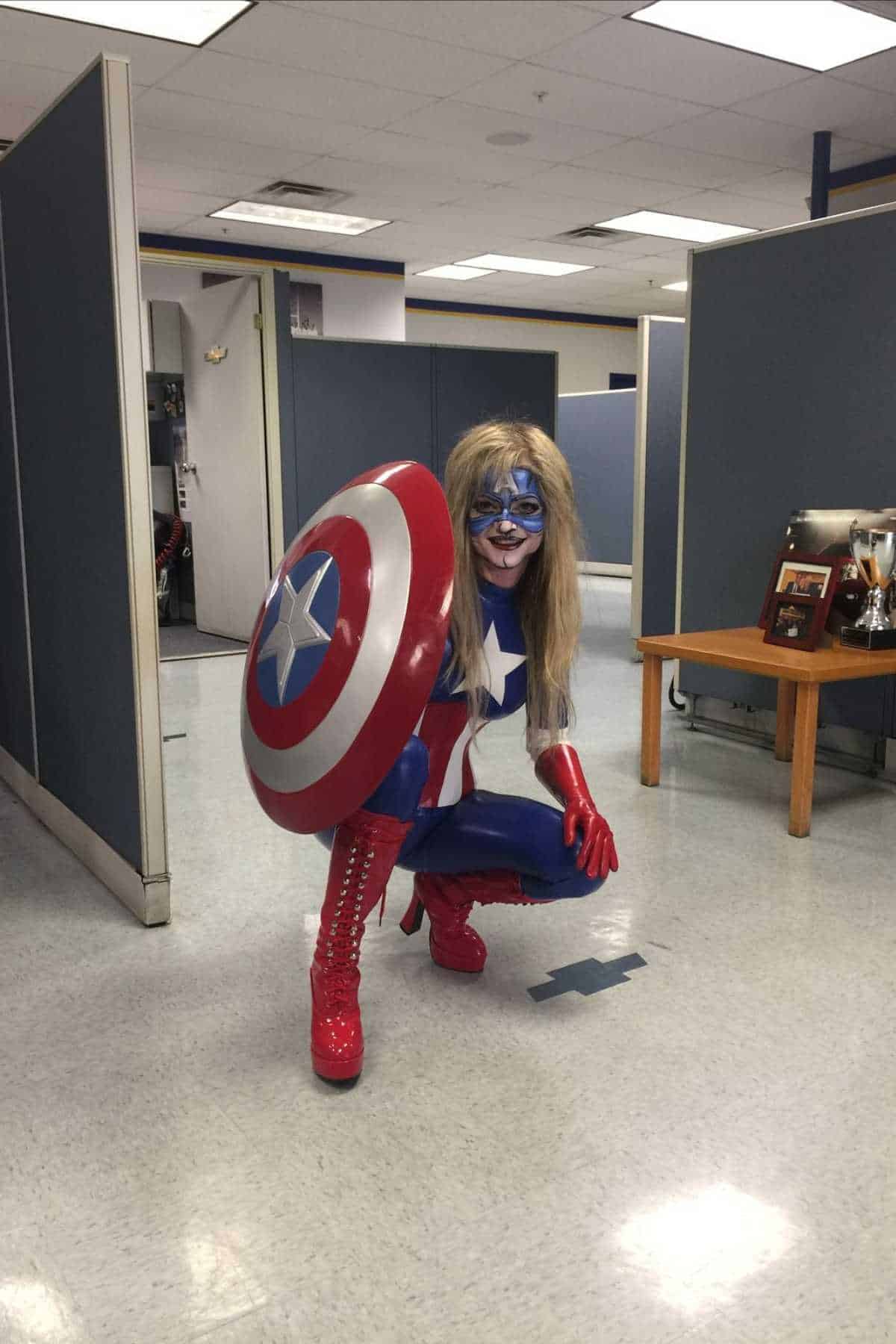 Person in a red, white, and blue superhero costume with a star shield crouches in an office with cubicles and a small table.