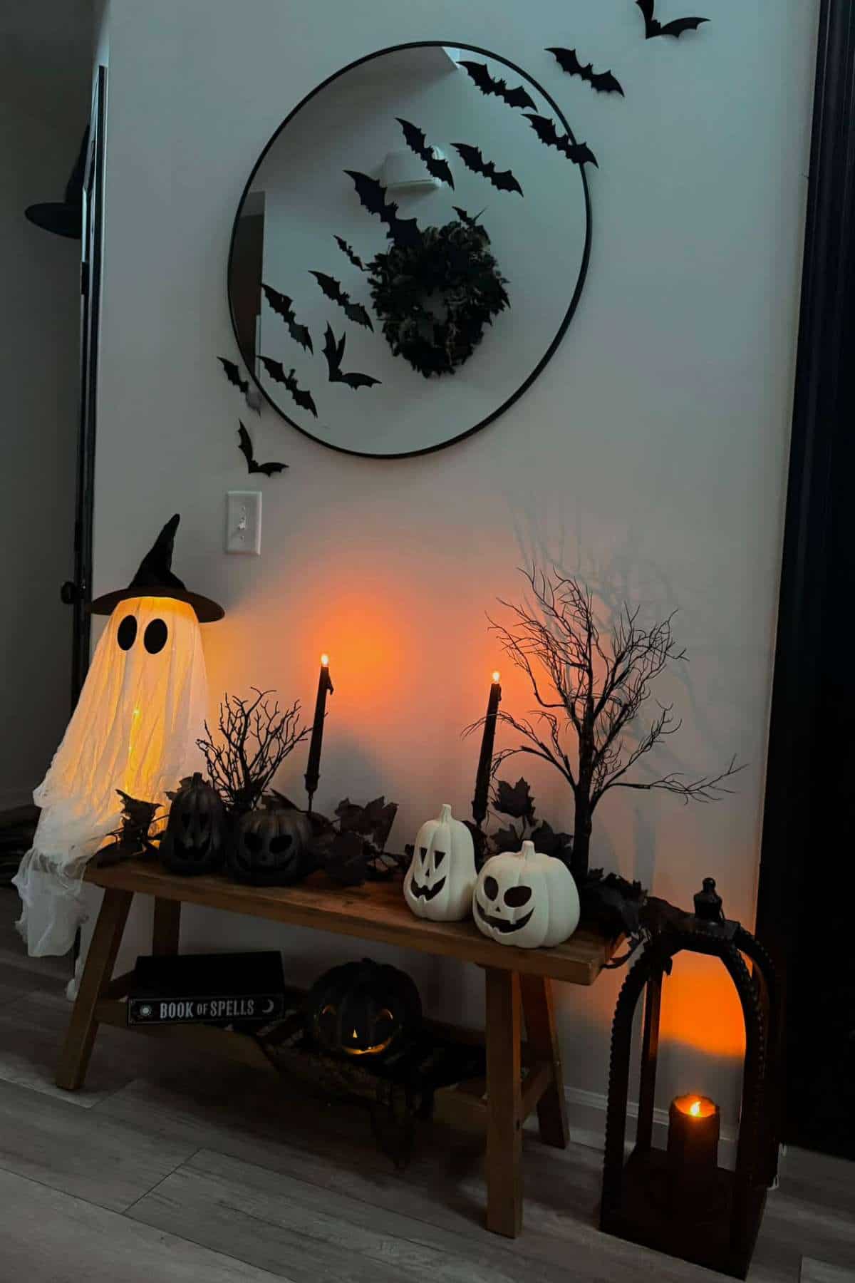 Halloween decorations include white pumpkins, a ghost, black trees, candles, bats on a mirror, and a book labeled "Book of Spells" arranged on a bench against a wall.