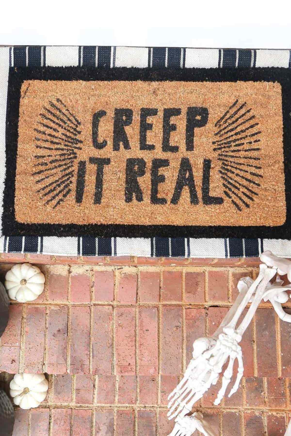 Doormat with the words "Creep It Real" in black lettering, placed on a striped rug next to small white pumpkins and a skeleton decoration.