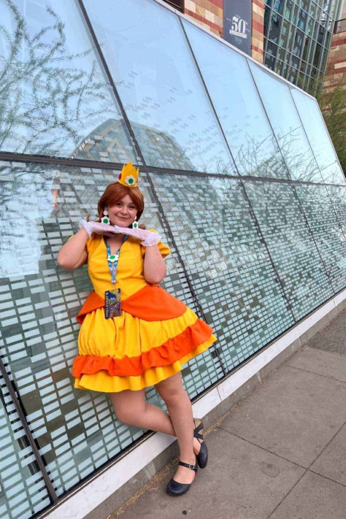 Person in a yellow and orange dress with a crown poses and smiles outside a building with glass windows and a brick façade.