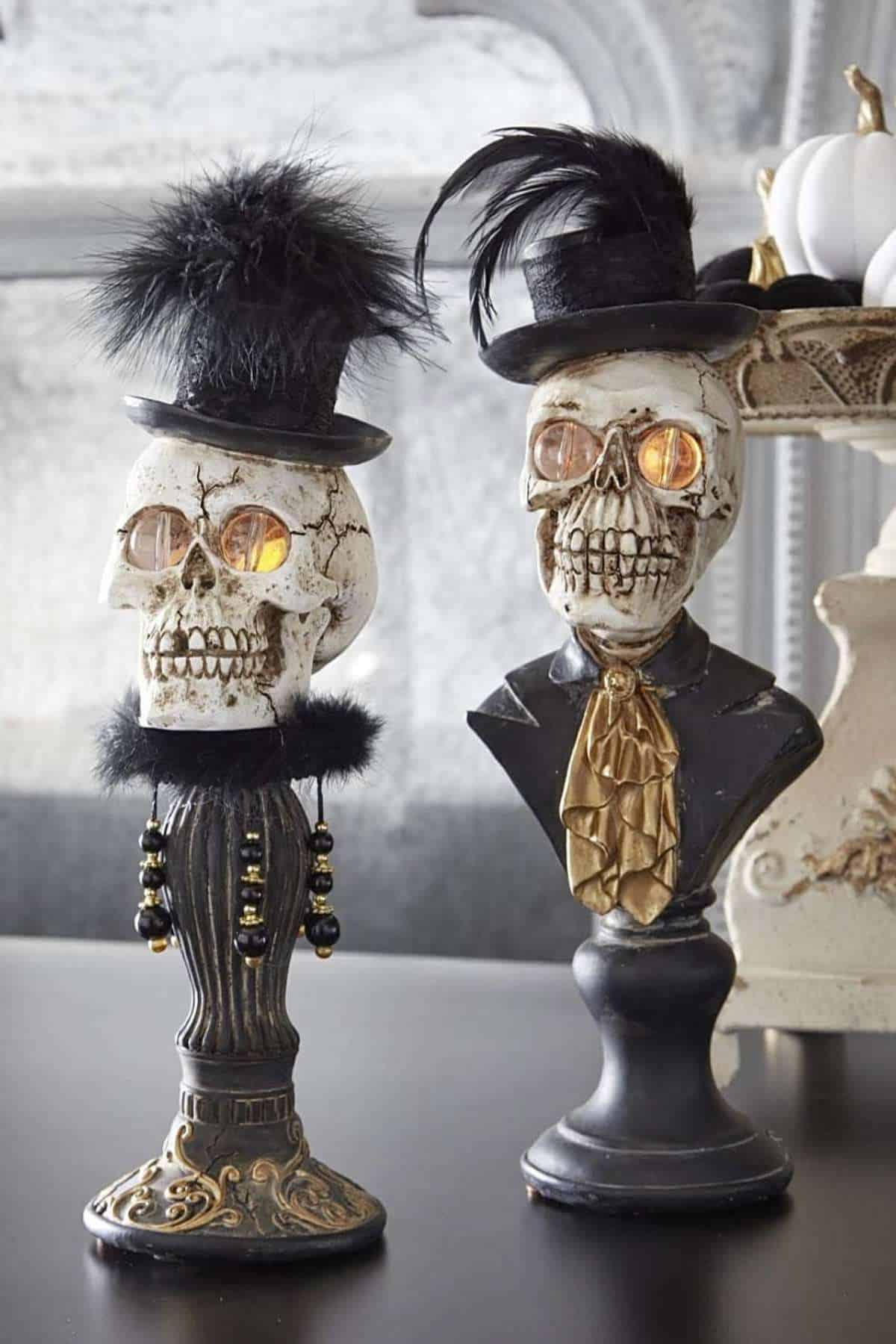 Two decorative skull busts wear top hats and formal attire, with glowing eyes and feather embellishments, displayed on a table in a gothic-inspired setting.