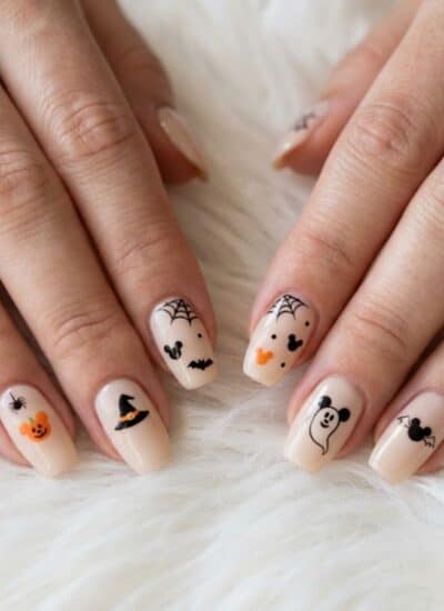 Close-up of hands with Halloween-themed nail art, including pumpkins, bats, spider webs, ghosts, a witch hat, and a bear, on a light base color.