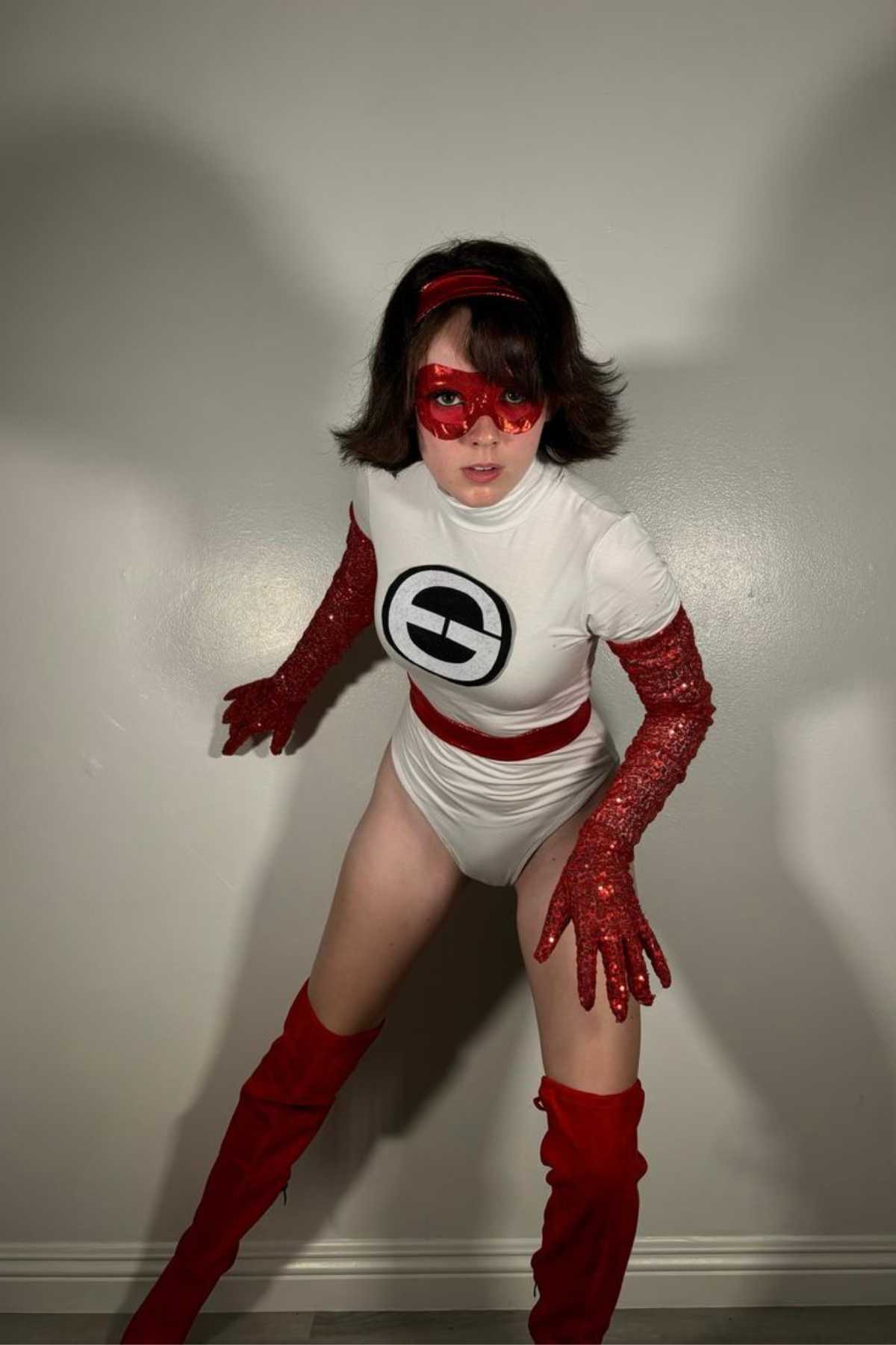 Person in a white superhero costume with a “G” logo, red gloves, red mask, red belt, and red boots, standing against a plain light-colored wall.