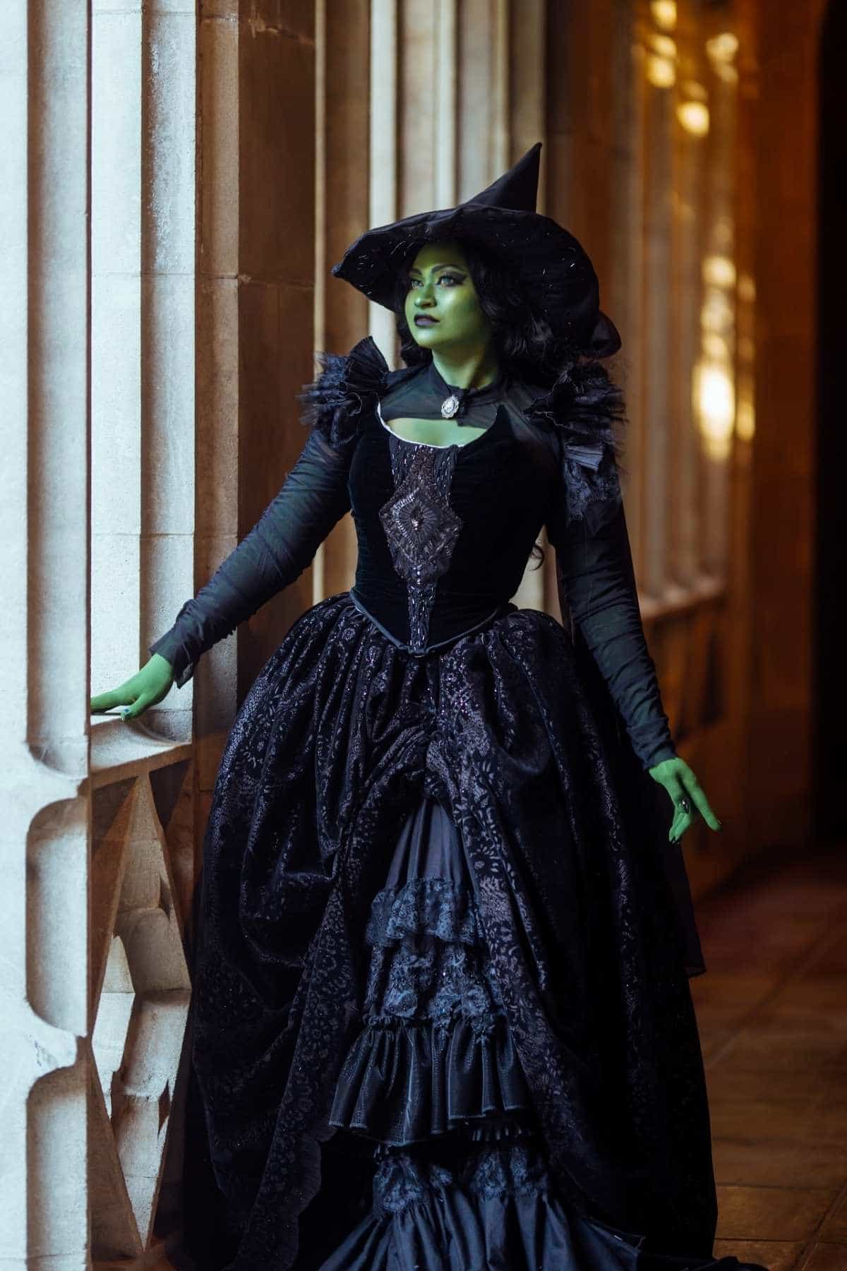 A person in a long black dress and pointed hat with green-painted skin stands by a stone window, dressed as a witch.