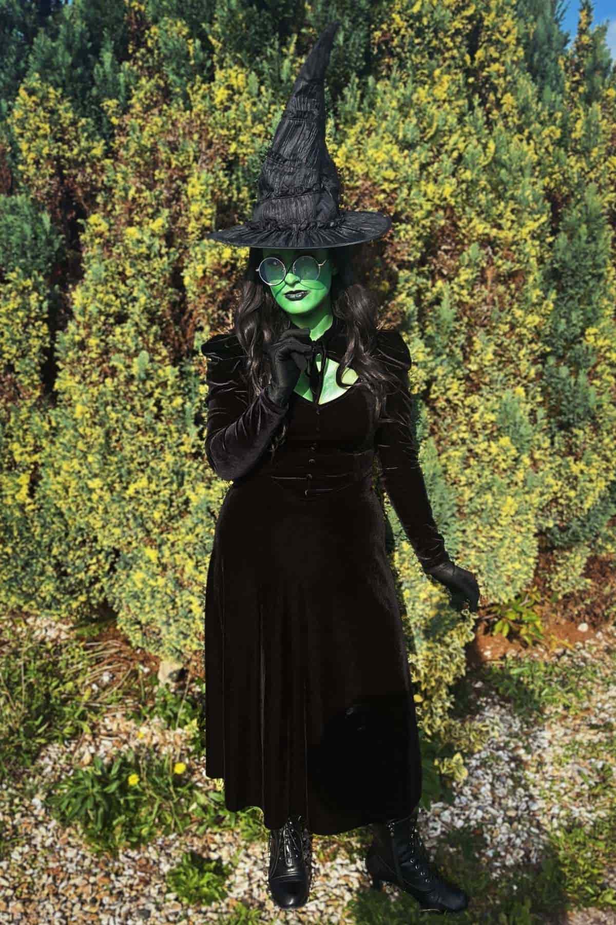 A person dressed as a witch with green face paint, a black hat, glasses, black dress, and boots stands outdoors in front of tall green bushes.