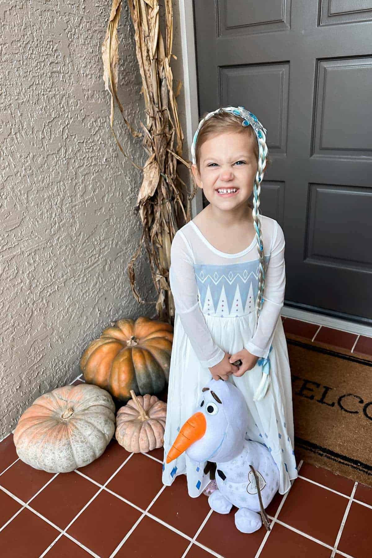 Young child in an Elsa costume stands on a porch with pumpkins, smiling and holding a plush Olaf toy, near a "welcome" mat and dried corn stalks.