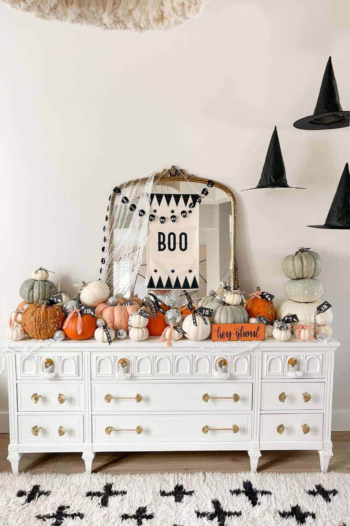 A white dresser decorated with assorted pumpkins, a "BOO" banner on the mirror, black garlands, and hanging witch hats, creating a Halloween-themed display.