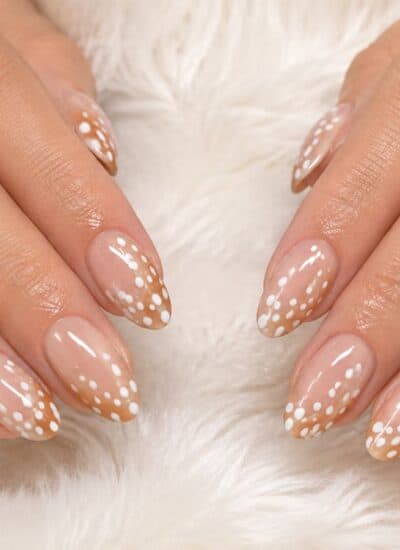 Two hands with almond-shaped nails featuring a nude base and white polka dot design, displayed on a white furry surface. The person is wearing gold rings.