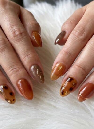 Two hands with almond-shaped nails featuring brown, amber, and taupe shades, some with gold accents and tortoiseshell patterns, resting on a white furry surface.