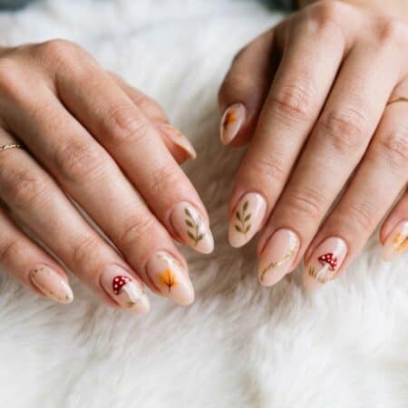 Hands with almond-shaped nails featuring autumn-themed nail art, including leaves and mushrooms, rest on a white, fuzzy surface.
