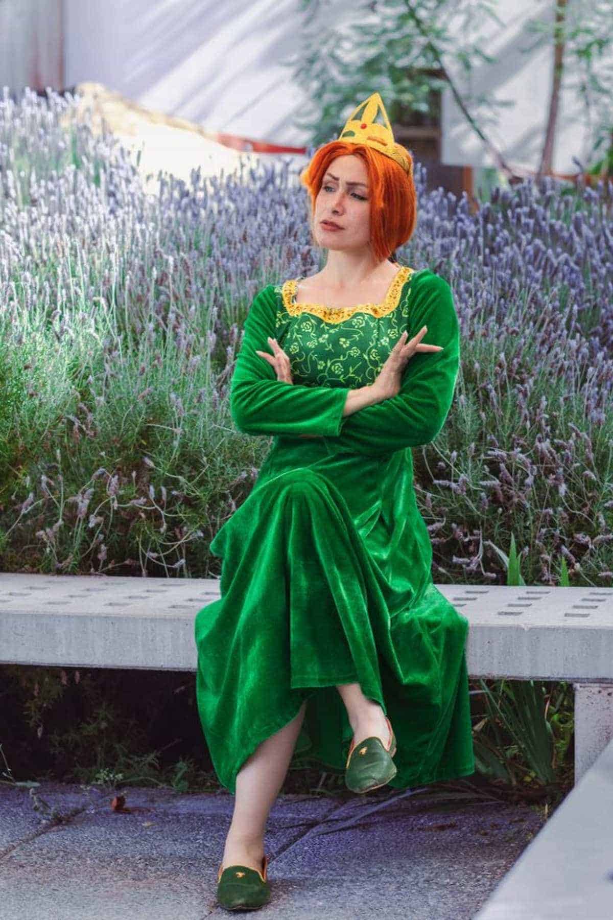 Person in a green medieval-style dress and crown sits on a bench outdoors, arms crossed, with a serious expression. Lavender plants are visible in the background.