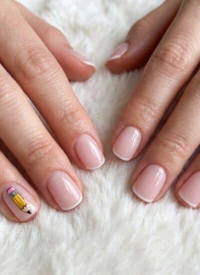 Two hands with short, natural-shaped nails painted in light pink; the left pinky has a pencil design, and the right ring finger has a small gold stud. Hands rest on a white textured surface.