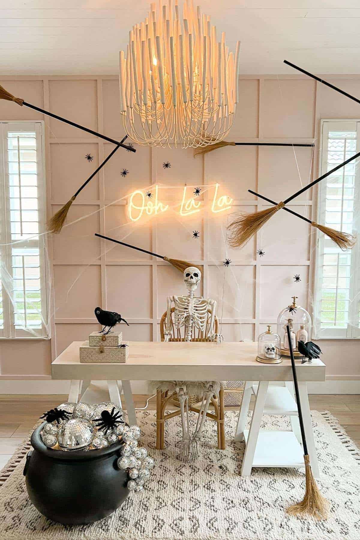 Skeleton seated at a decorated desk with broomsticks, spiders, a cauldron, and a neon "Ooh la la" sign in a Halloween-themed room.
