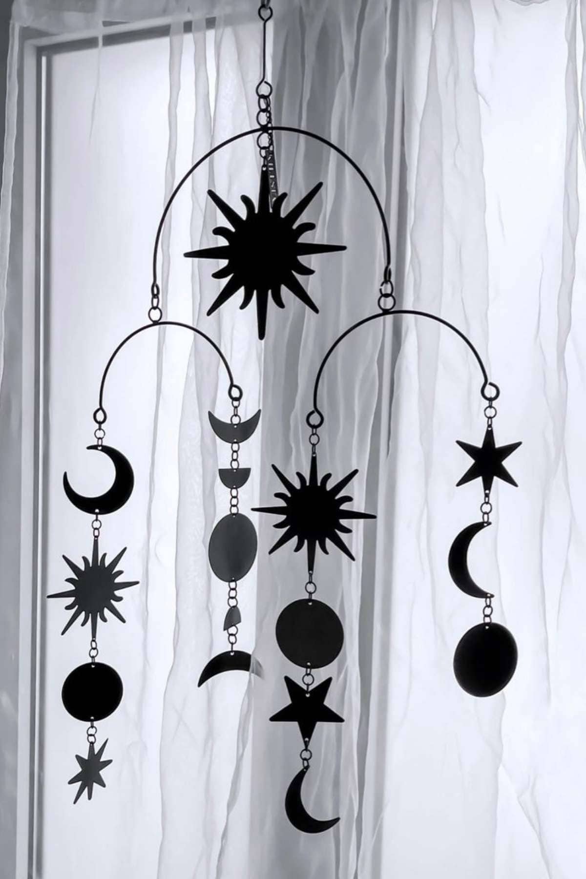 A metal mobile with star, moon, and sun shapes hangs in front of a sheer curtain and window.
