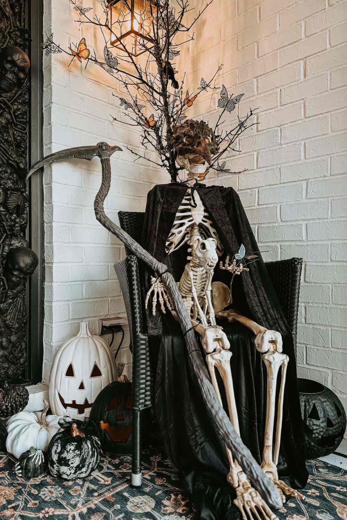 A skeleton in a black cloak sits on a chair holding a scythe, surrounded by pumpkins and Halloween decorations against a white brick wall.