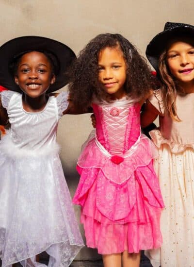 Five children in costumes stand in a row against a plain wall, smiling. Outfits include a knight, witch, princess, fairy, and ladybug. Some hold small containers for treats.