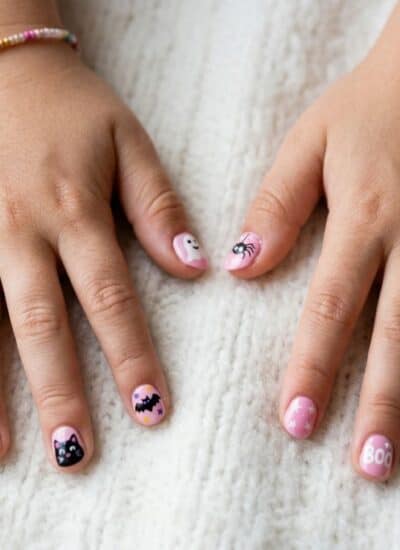 Child’s hands on a white blanket, nails painted with Halloween designs like pumpkins, bats, spiders, and the word “Boo.” Wears a beaded bracelet with a star charm.