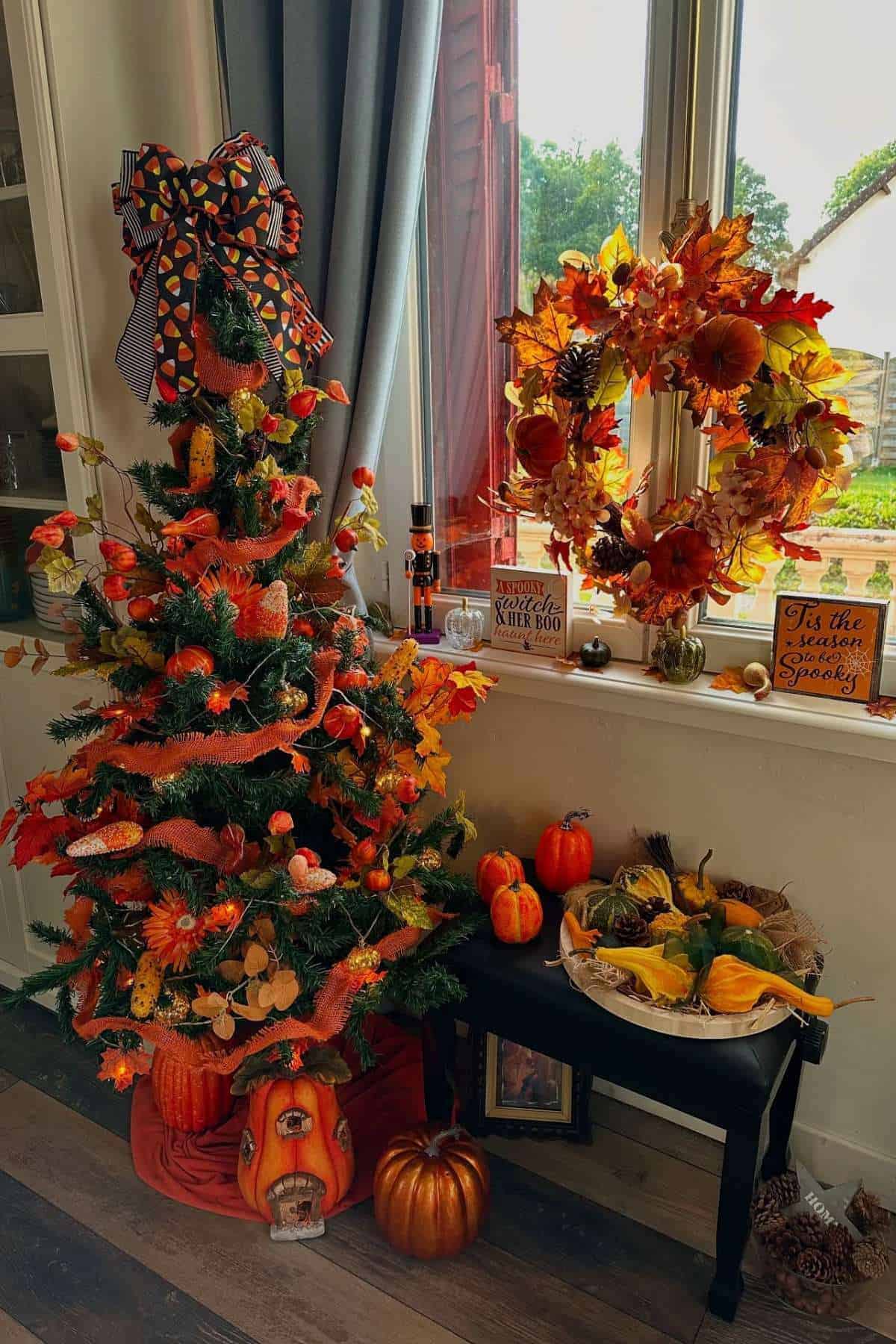 A small Christmas tree and a wreath decorated with autumn leaves, pumpkins, and gourds, arranged by a window with seasonal decor on a black bench and floor.