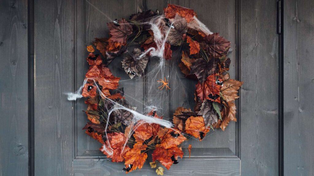 A wreath made of orange and brown autumn leaves hangs on a door, decorated with fake spider webs and plastic spiders for Halloween.