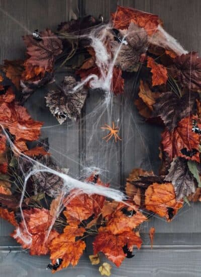 A wreath made of orange and brown autumn leaves hangs on a door, decorated with fake spider webs and plastic spiders for Halloween.