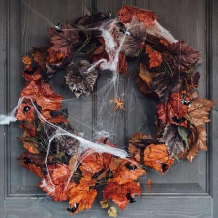 A wreath made of orange and brown autumn leaves hangs on a door, decorated with fake spider webs and plastic spiders for Halloween.