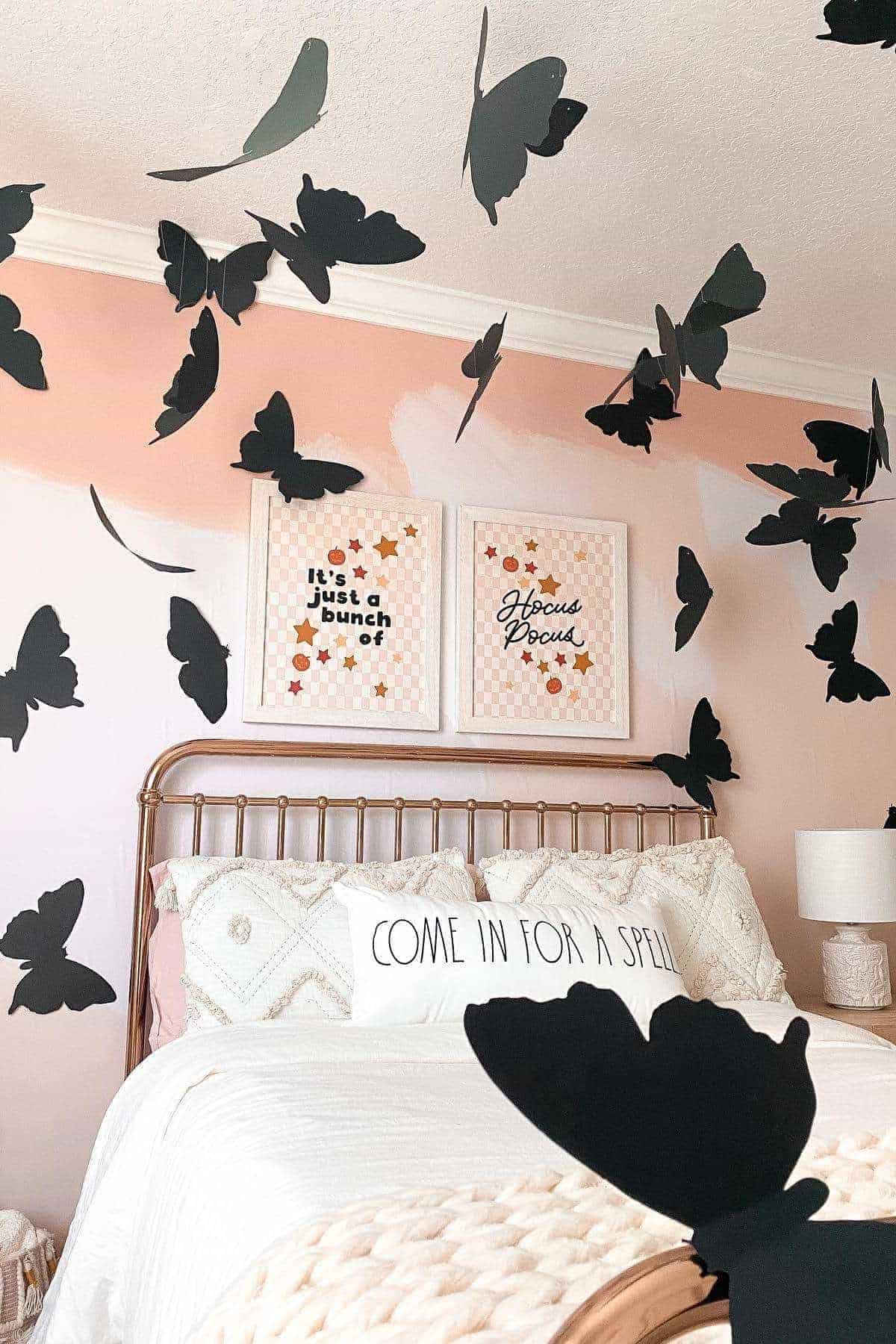 A bedroom with a white bed, decorative pillows, two framed prints on the wall, and large black butterfly cutouts attached to the wall and ceiling.