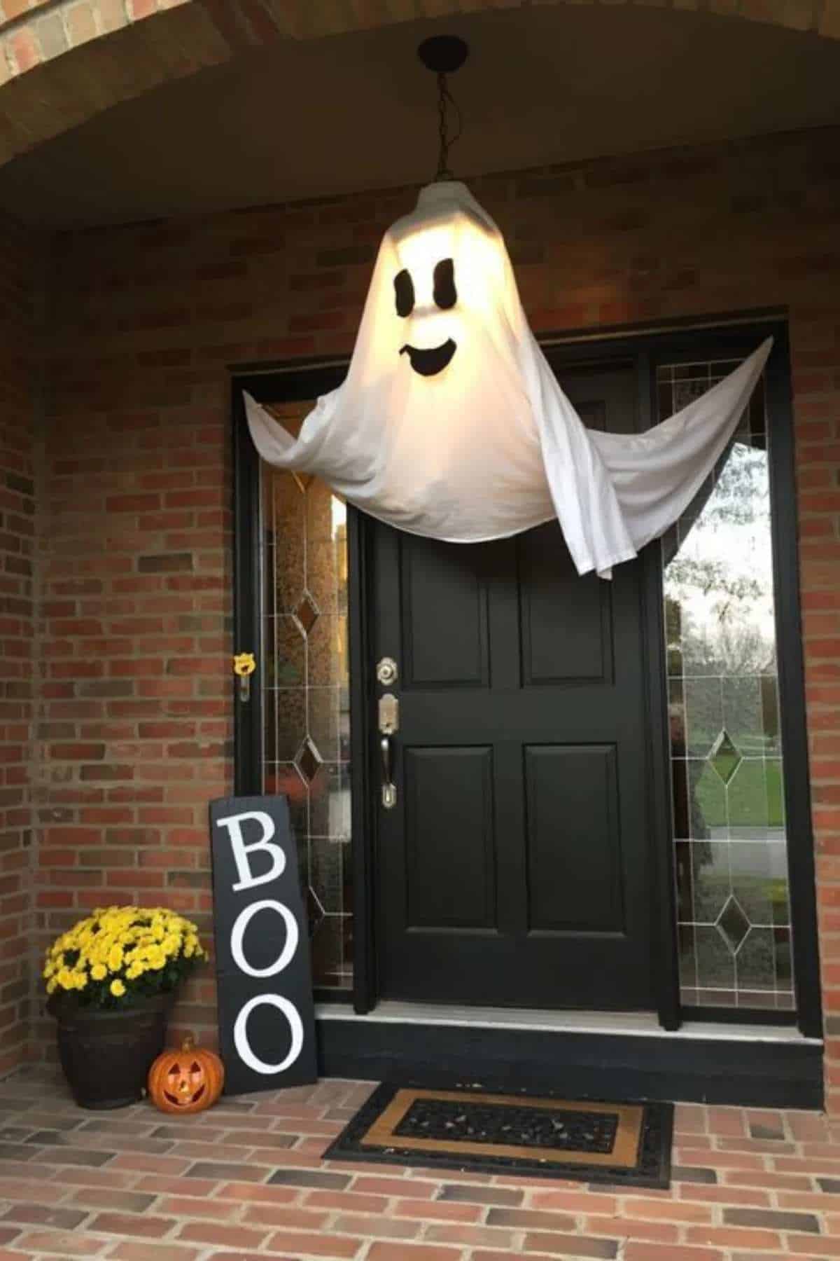 A large ghost decoration hangs over a black front door with a "BOO" sign, potted yellow flowers, and a carved pumpkin on the brick porch.