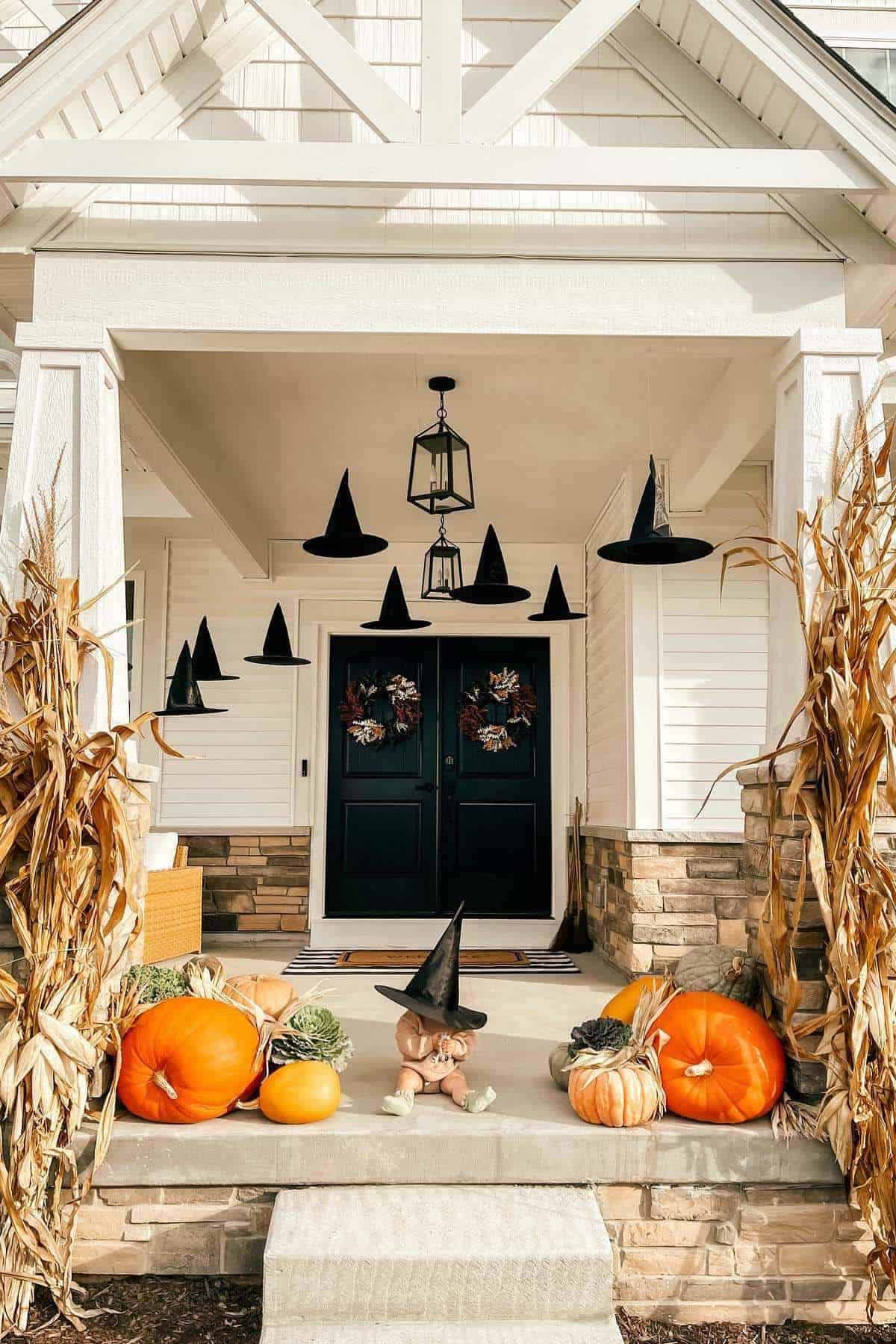 Front porch decorated with pumpkins, gourds, dried cornstalks, hanging witch hats, and a teddy bear wearing a witch hat in front of double black doors.