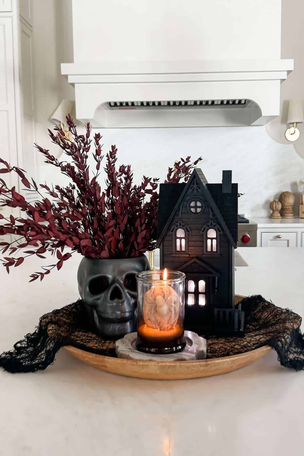 A decorative tray with a black skull, burgundy foliage, a lit candle, and a miniature haunted house sits on a kitchen island.