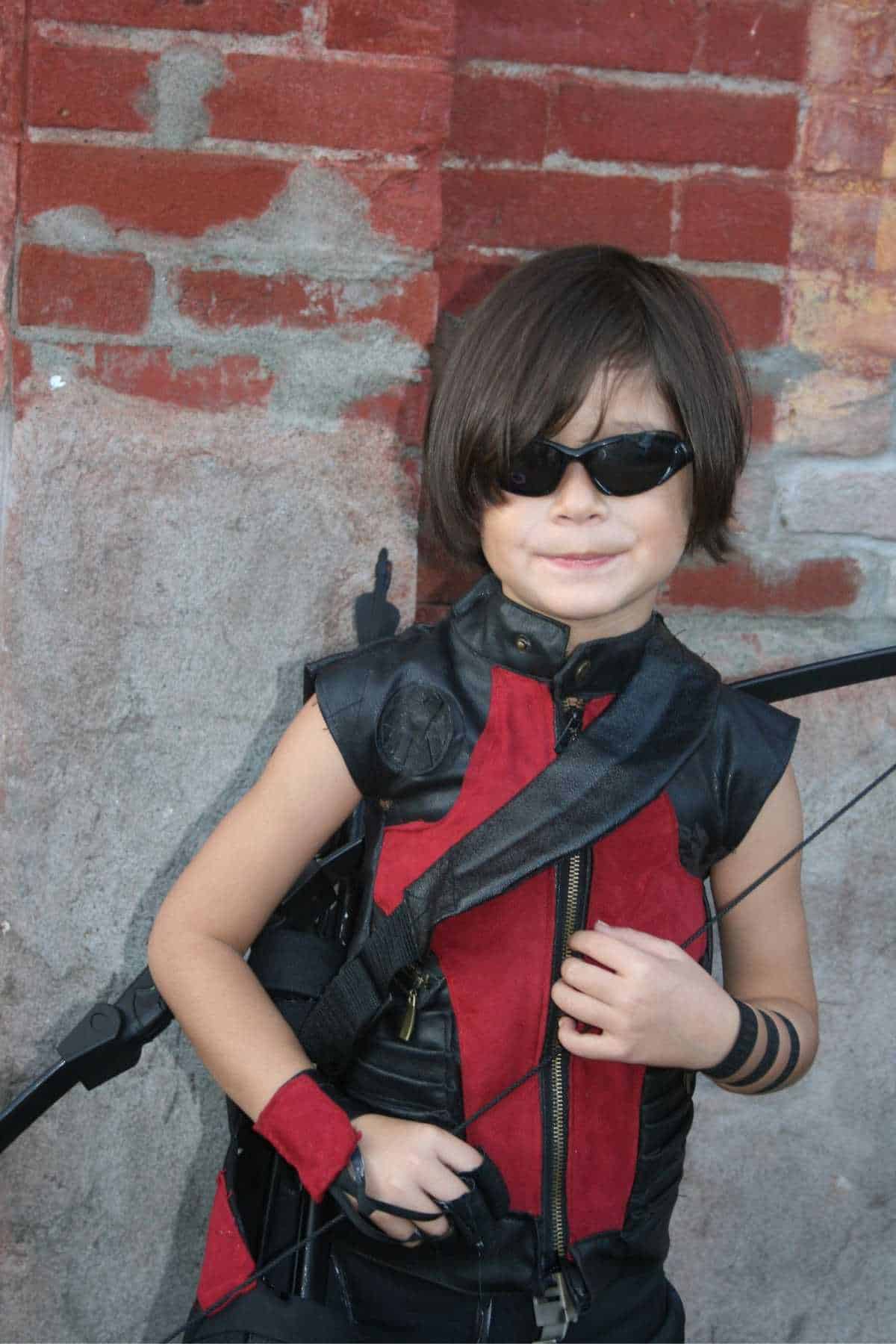 Child wearing a red and black costume, sunglasses, and fingerless gloves, standing against a brick wall and holding a bow.